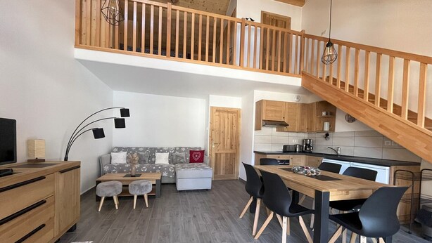 Un apartamento moderno estilo loft con una escalera de madera, un área de comedor con una mesa y sillas de madera, una cocina con armarios de madera y una zona de estar acogedora con un sofá y otomanas.