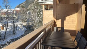 Un balcón de madera con una mesa y sillas, con vista a un paisaje nevado que incluye un río y edificios al fondo.