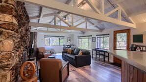 A spacious living area with a stone wall, a wooden ceiling, and a large window. - Waterfall House - Riverfront - BBQ - Sleeps 4 (Oakhurst)