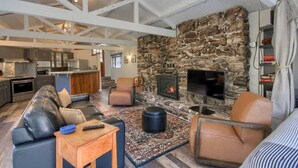 A spacious living area with a stone fireplace, leather sofas, and a kitchen in the background. - Waterfall House - Riverfront - BBQ - Sleeps 4 (Oakhurst)