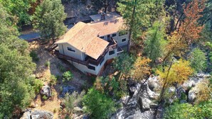 A two-story house with a metal roof, surrounded by trees and a rocky stream. - Waterfall House - Riverfront - BBQ - Sleeps 4 (Oakhurst)