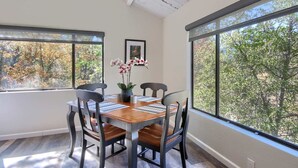 A dining area with a wooden table and chairs, a vase with flowers, and large windows with a view of trees. - Waterfall House - Riverfront - BBQ - Sleeps 4 (Oakhurst)