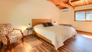 Une chambre avec un plafond en bois, un lit avec une literie blanche, un fauteuil à motifs et une petite table de chevet avec une lampe.
