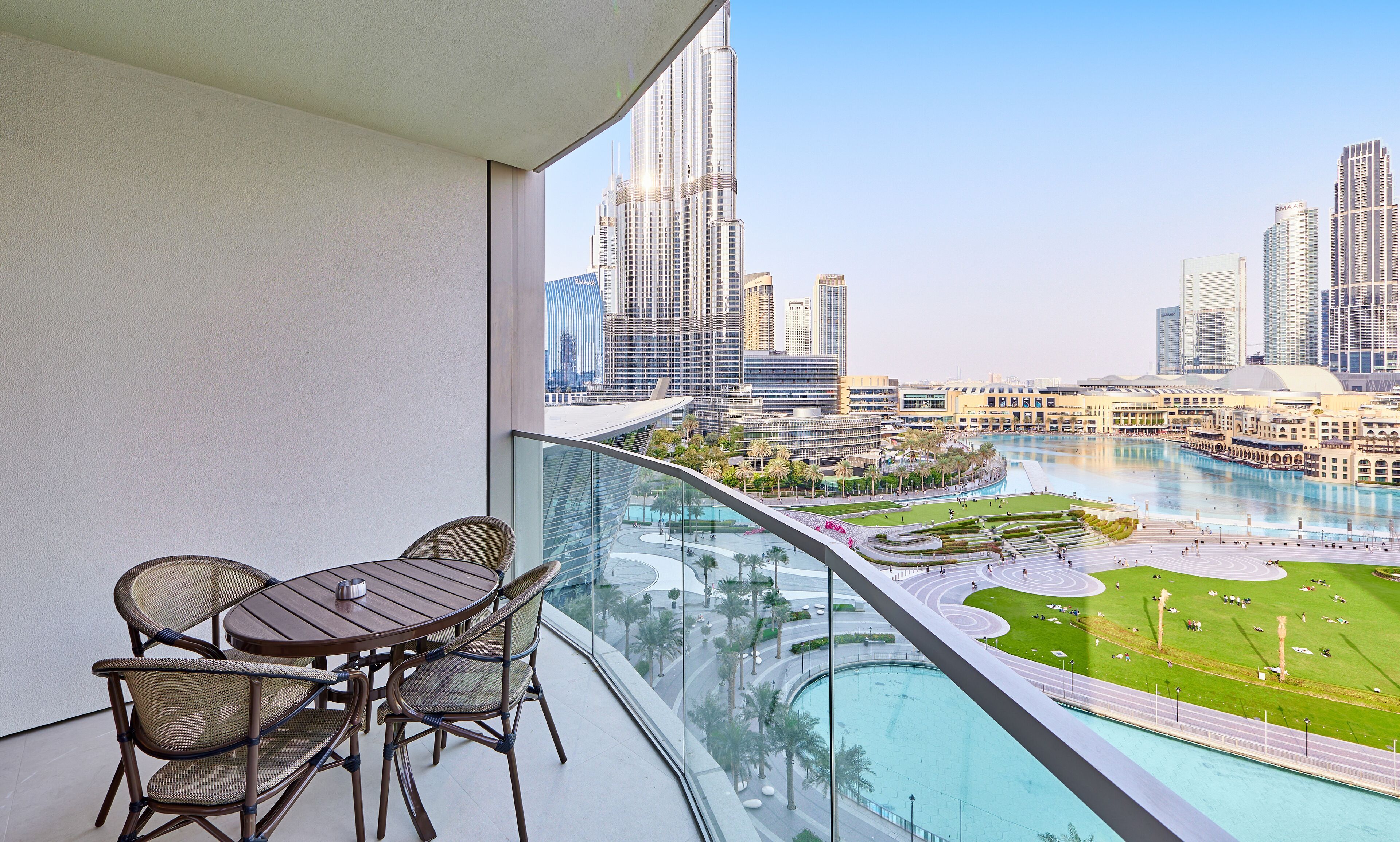 A balcony with a table and chairs, overlooking a cityscape with tall buildings and a water feature.