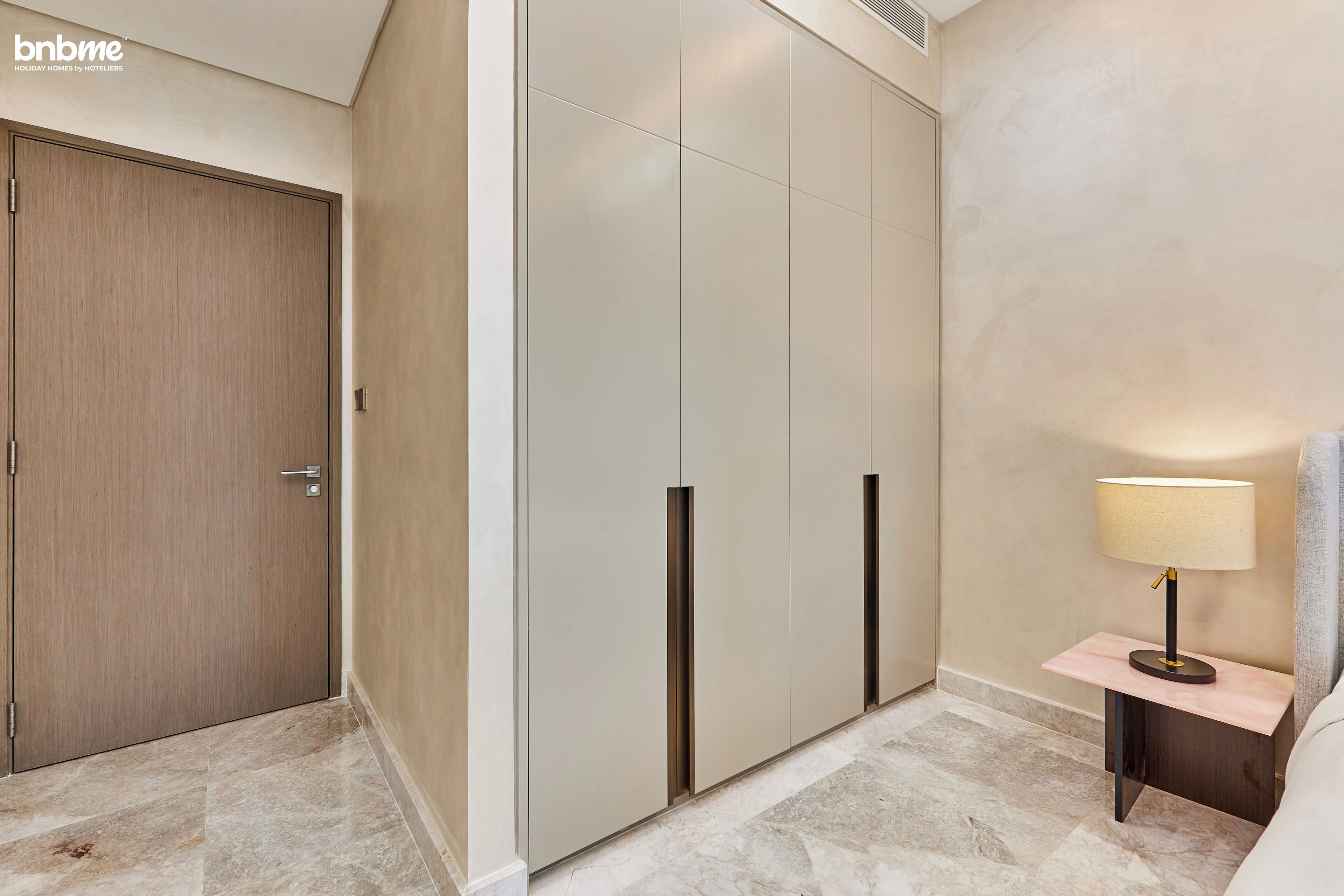 A hotel room with a wooden door, a large wardrobe, a bedside table with a lamp, and marble flooring.