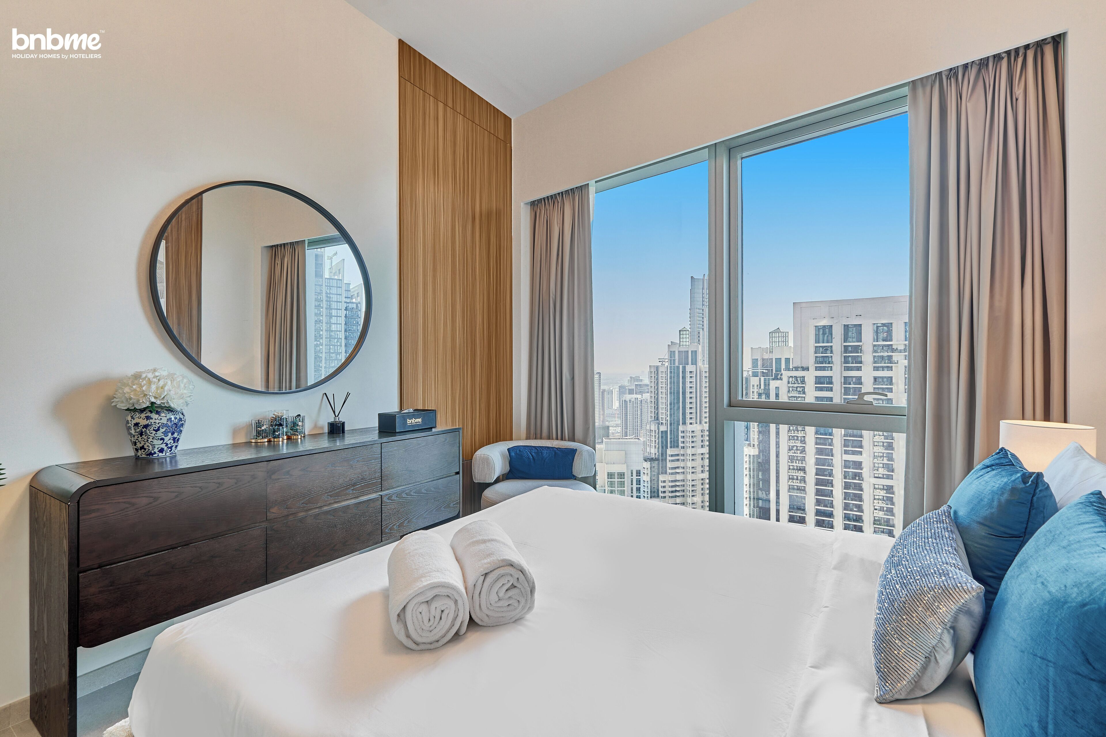A hotel room with a large bed, a wooden dresser, a round mirror, and a view of the cityscape through large windows.
