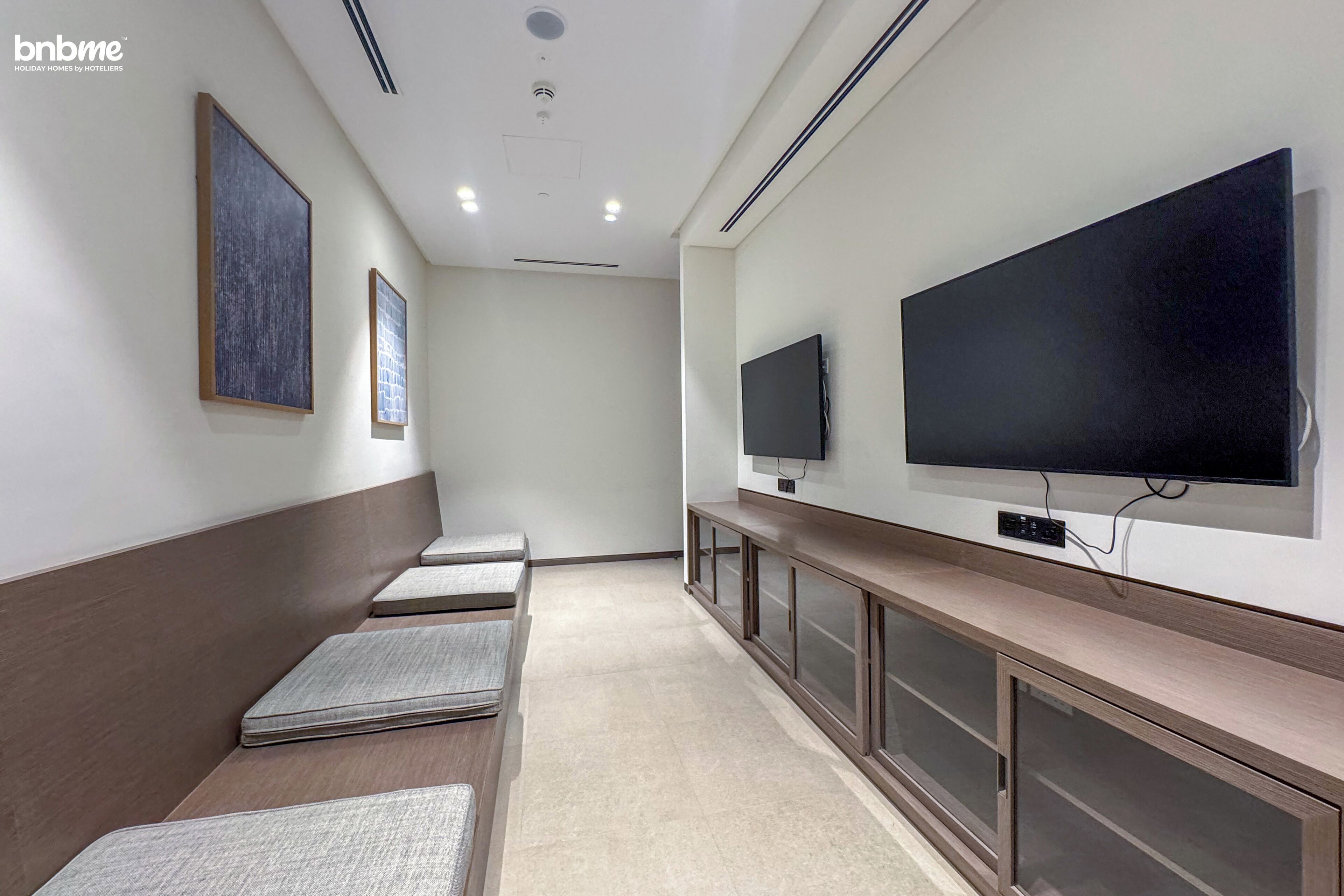 A hotel lobby with a long bench seating area, a large flat-screen TV mounted on the wall, and framed artwork on the walls.