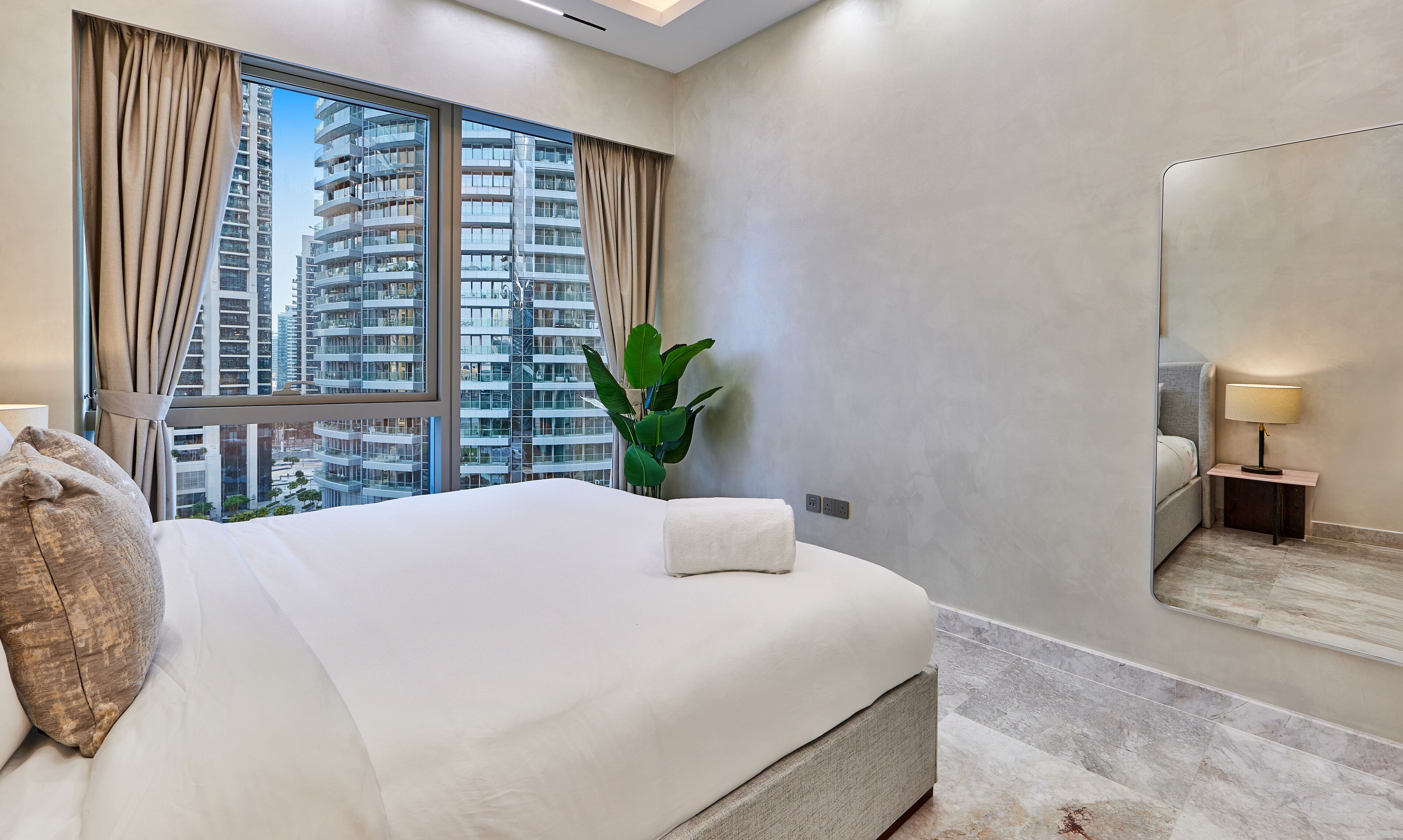A hotel room with a large bed, a nightstand with a lamp, a potted plant, and a view of high-rise buildings.