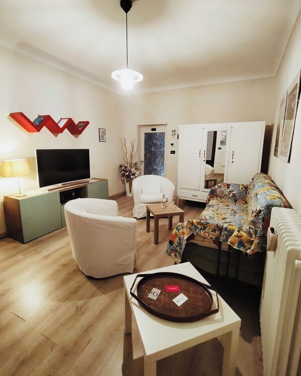 Ein modernes Wohnzimmer mit Flachbildfernseher, einem weißen Sessel, einem Sofa mit bunter Decke, einem kleinen Tisch und einer Wanddekoration.