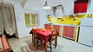A compact kitchen with a dining area, featuring a red tablecloth, wooden chairs, a yellow backsplash, and a white refrigerator. - La Casetta sul Lago (Viverone)