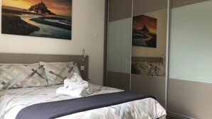 A bedroom with a bed, a large mirror, and a painting of a sunset over a castle. - Blackberry Cottage country cottage (Lessay)