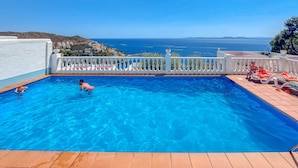A swimming pool with clear blue water, a white fence, and a view of the sea and hills in the background. - House in Rosas With Waterfront & Beach Nearby (Roses)
