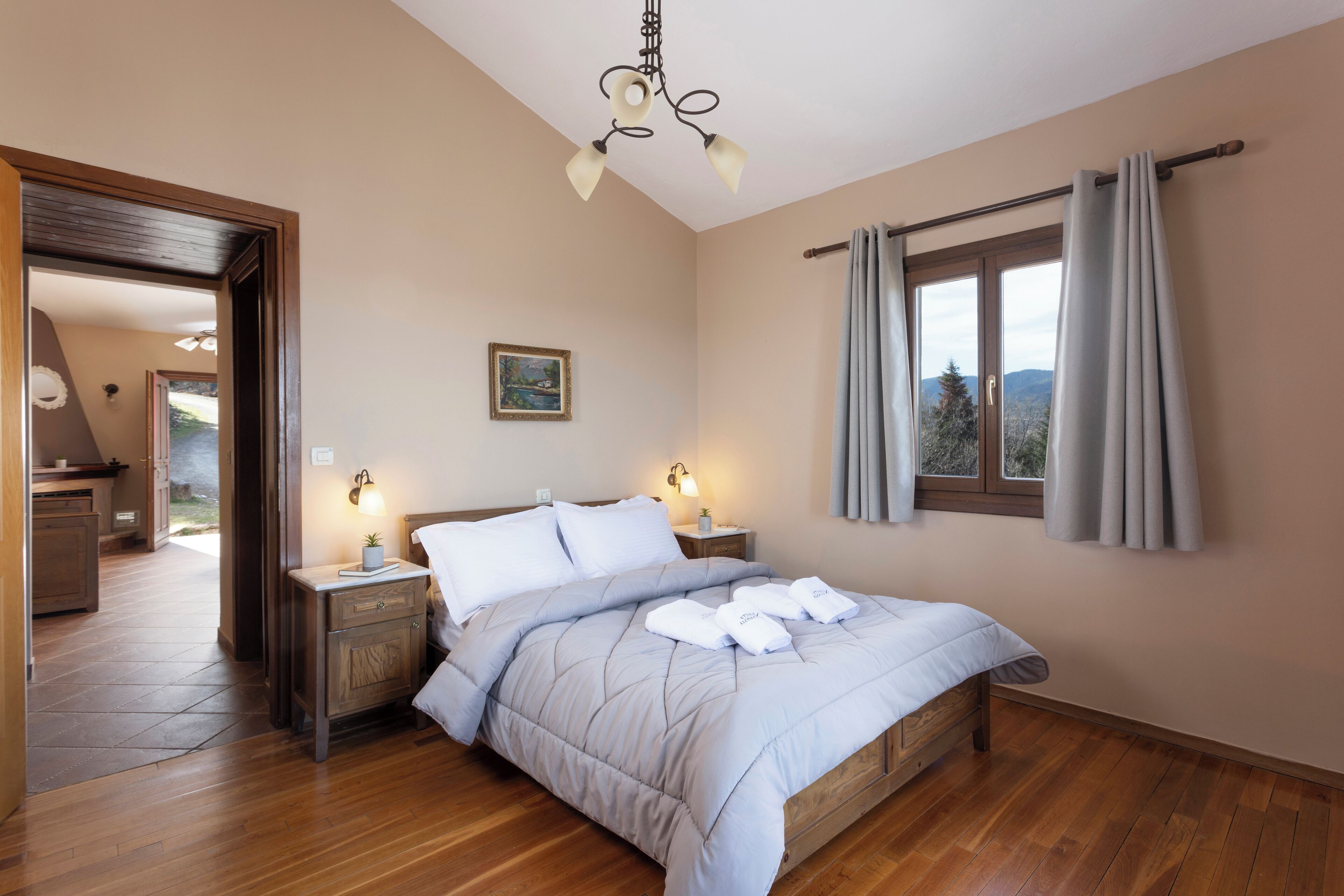 A bedroom with a wooden floor, a bed with a quilt, two bedside tables with lamps, a window with curtains, and a painting on the wall.