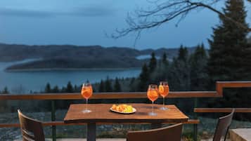 A table with two glasses of orange drink and a plate of fruit, set against a scenic view of a lake and mountains.