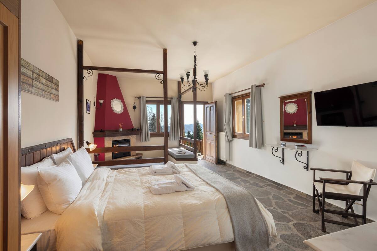 Ein Schlafzimmer mit einem großen Bett, Steinboden, Holzbalkendecke, einer Kristallleuchtre, einem Kamin und einem an der Wand befestigten Fernseher.