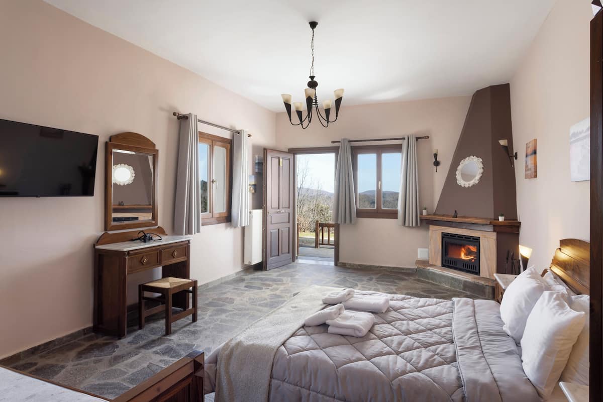 Ein Schlafzimmer mit Steinfußboden, einem Bett mit weißer Bettwäsche, einem Schminktisch, einem Spiegel, einem Fernseher, einem Kamin und Blick ins Freie.