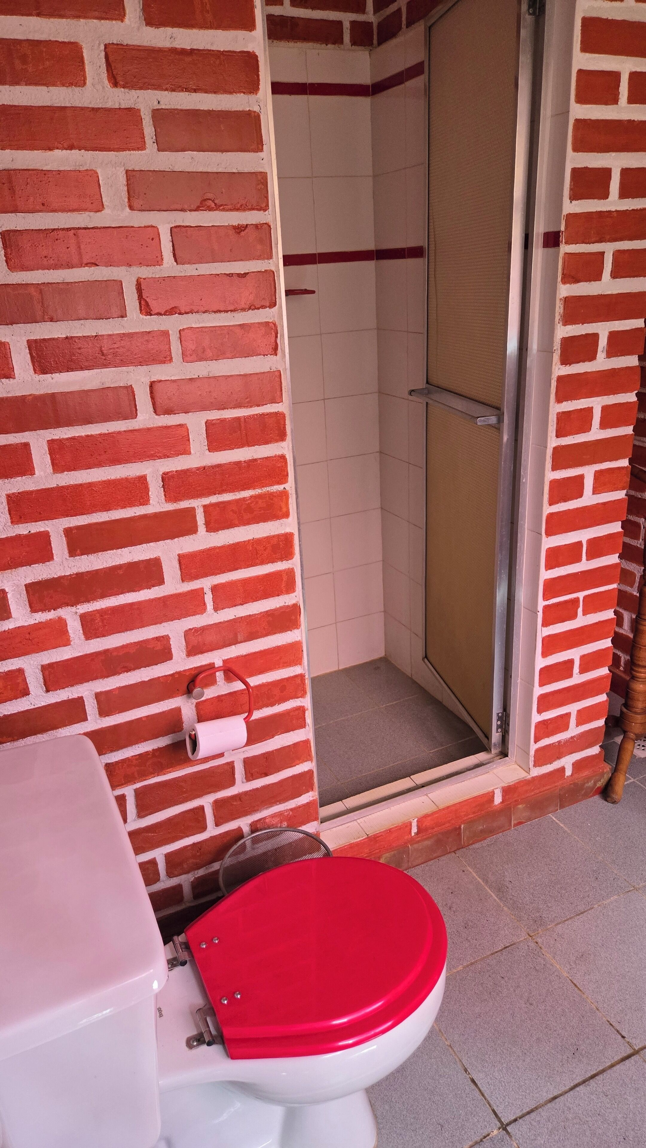 Un baño con inodoro rojo, ducha con puerta de cristal y pared de azulejos blancos.