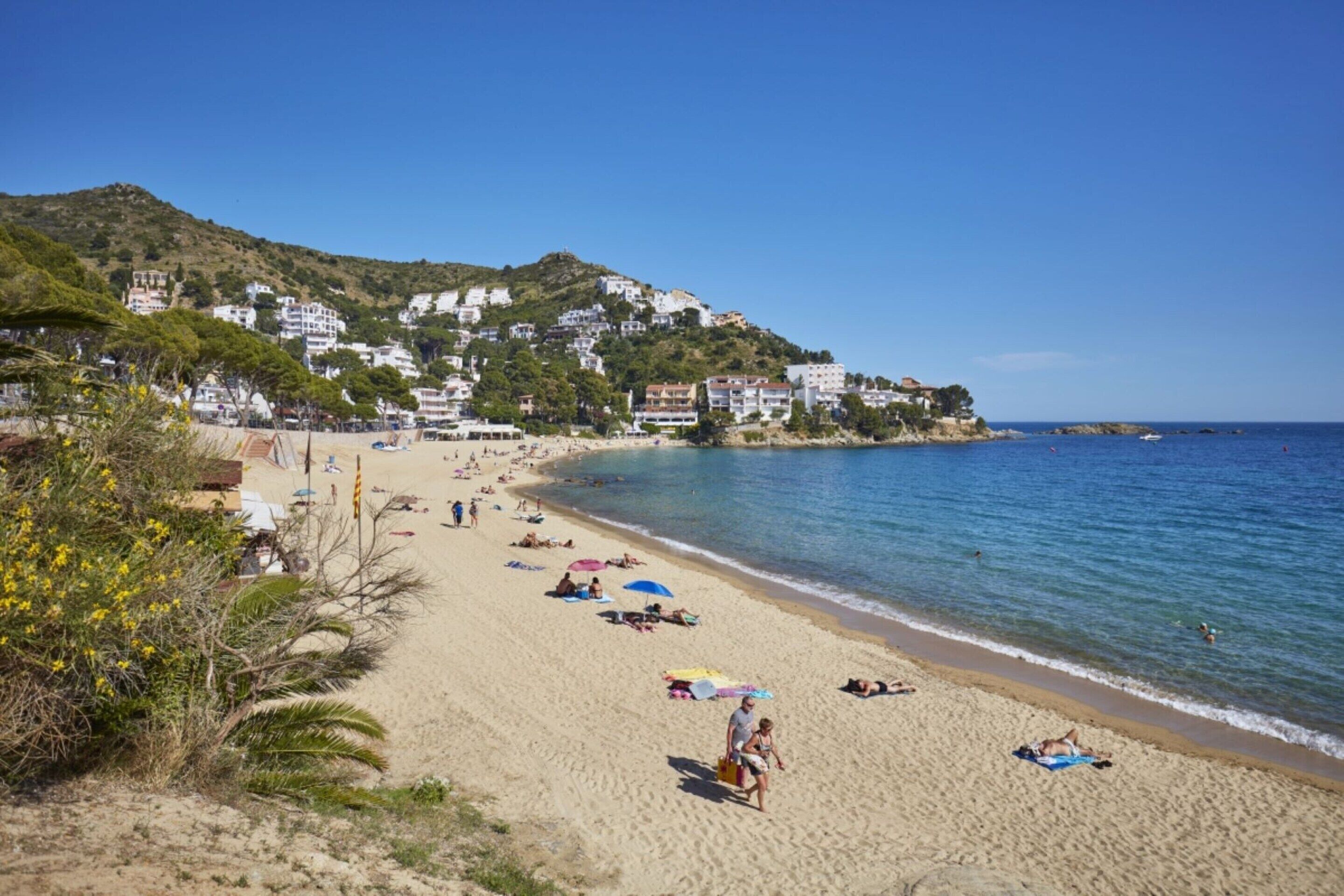 A beach with people sunbathing, umbrellas, and buildings in the background.