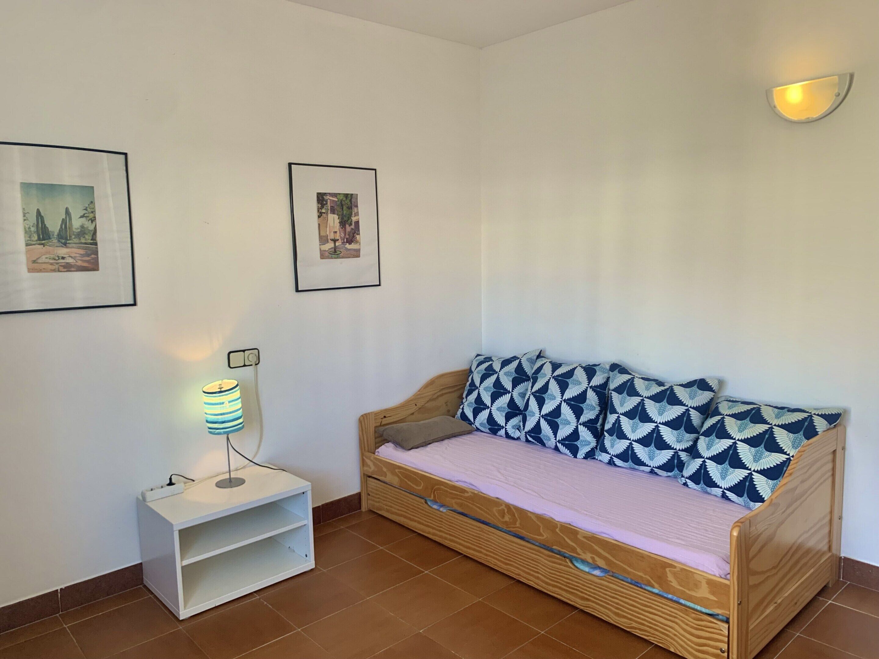A cozy room with a wooden sofa, a small white nightstand with a lamp, and two framed pictures on the wall.