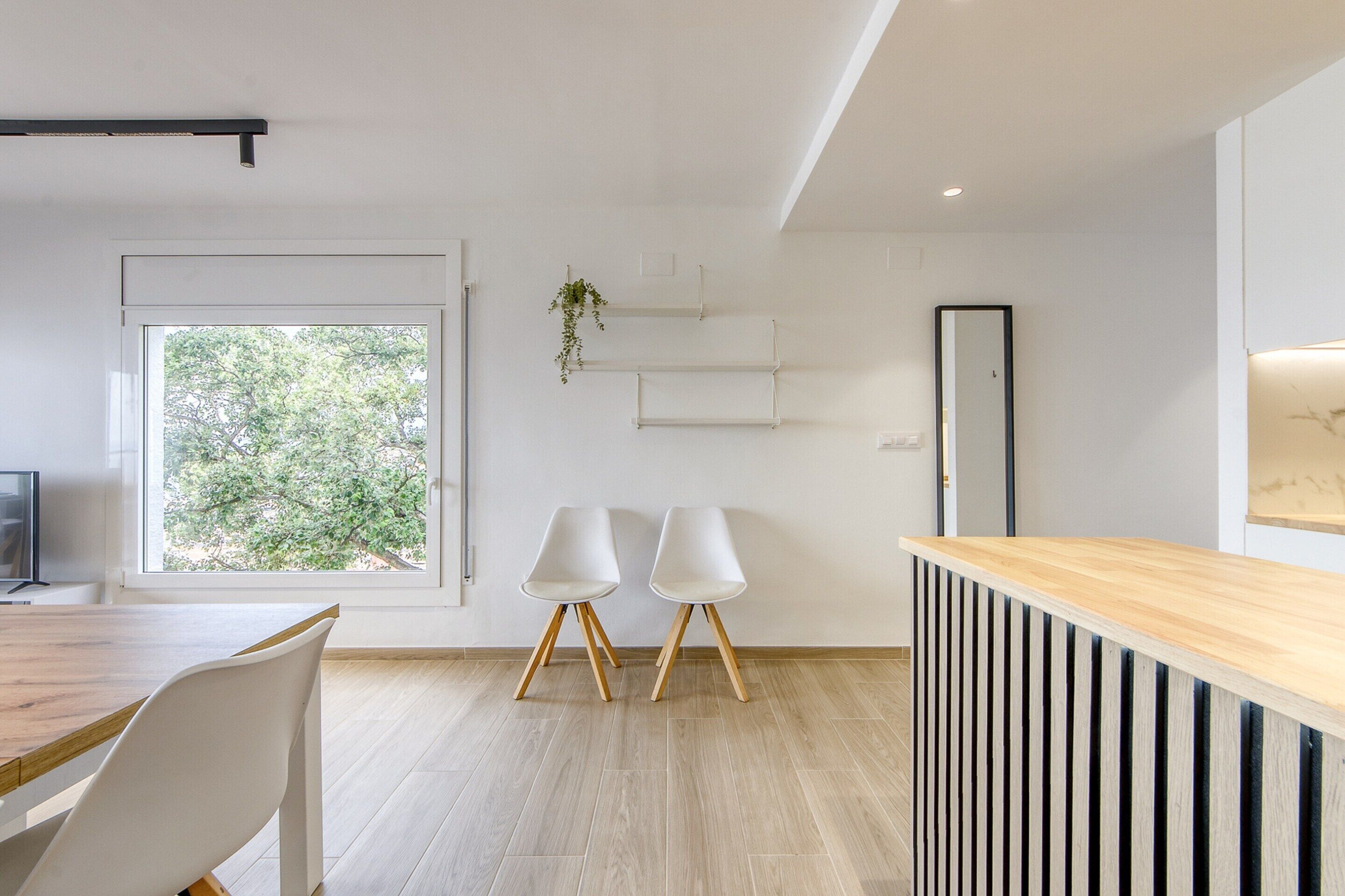 A modern, minimalist room with a wooden dining table, two white chairs, a wooden reception desk, and a large window offering a view of trees.