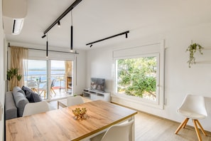 Ett modernt vardagsrum med ett matbord i trä, vita stolar, en soffa och en tv. Stora fönster ger utsikt över omgivningen.