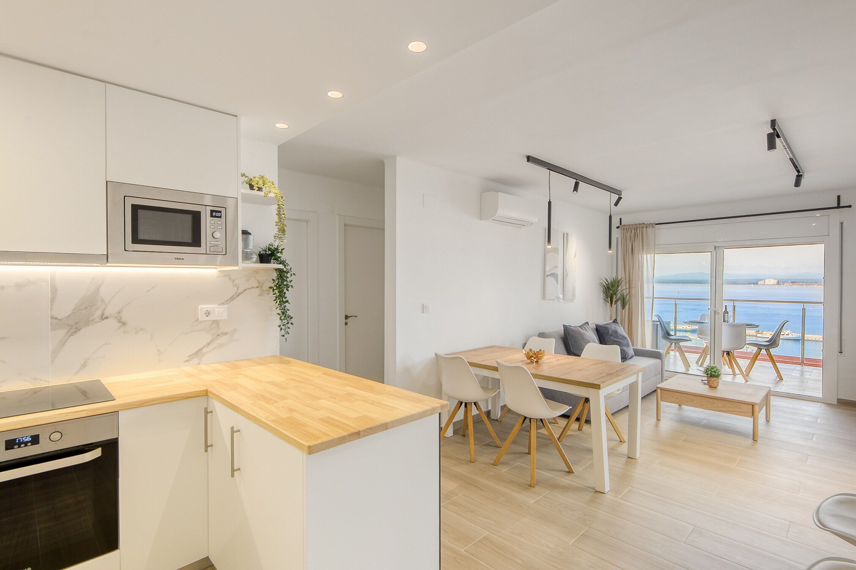 A modern kitchen with a built-in microwave, oven, and wooden countertop. There is a dining area with a table and chairs, and a sofa. The room has large windows with a view of the sea.