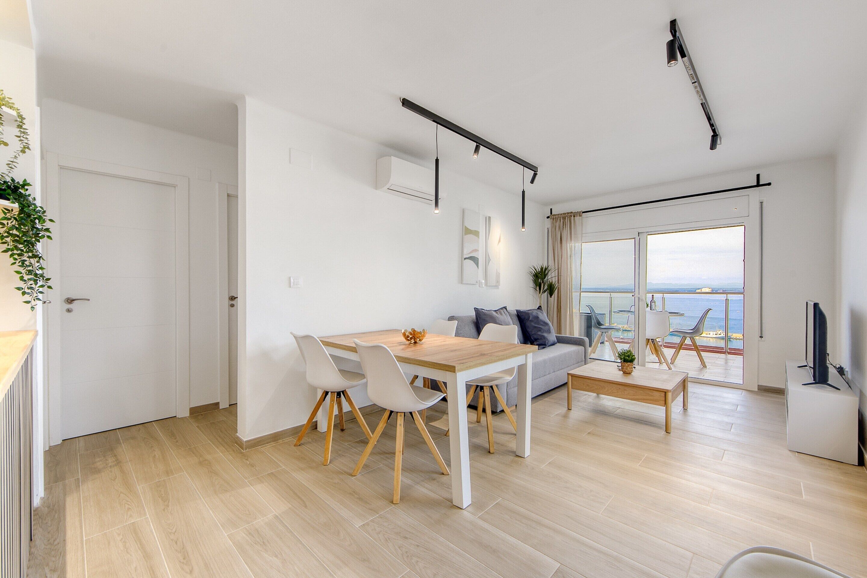 A modern living room with a wooden dining table, white chairs, a sofa, and a coffee table. There is a TV on a stand and a plant on the windowsill.