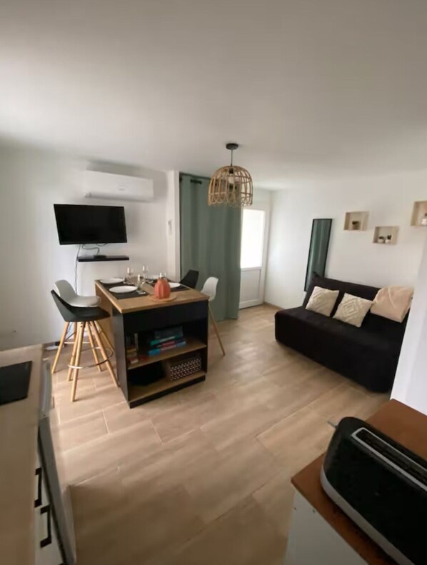 Un salon moderne avec un canapé noir, une table à manger en bois avec des chaises, un téléviseur fixé au mur et un coin cuisine équipé d’un grille-pain.