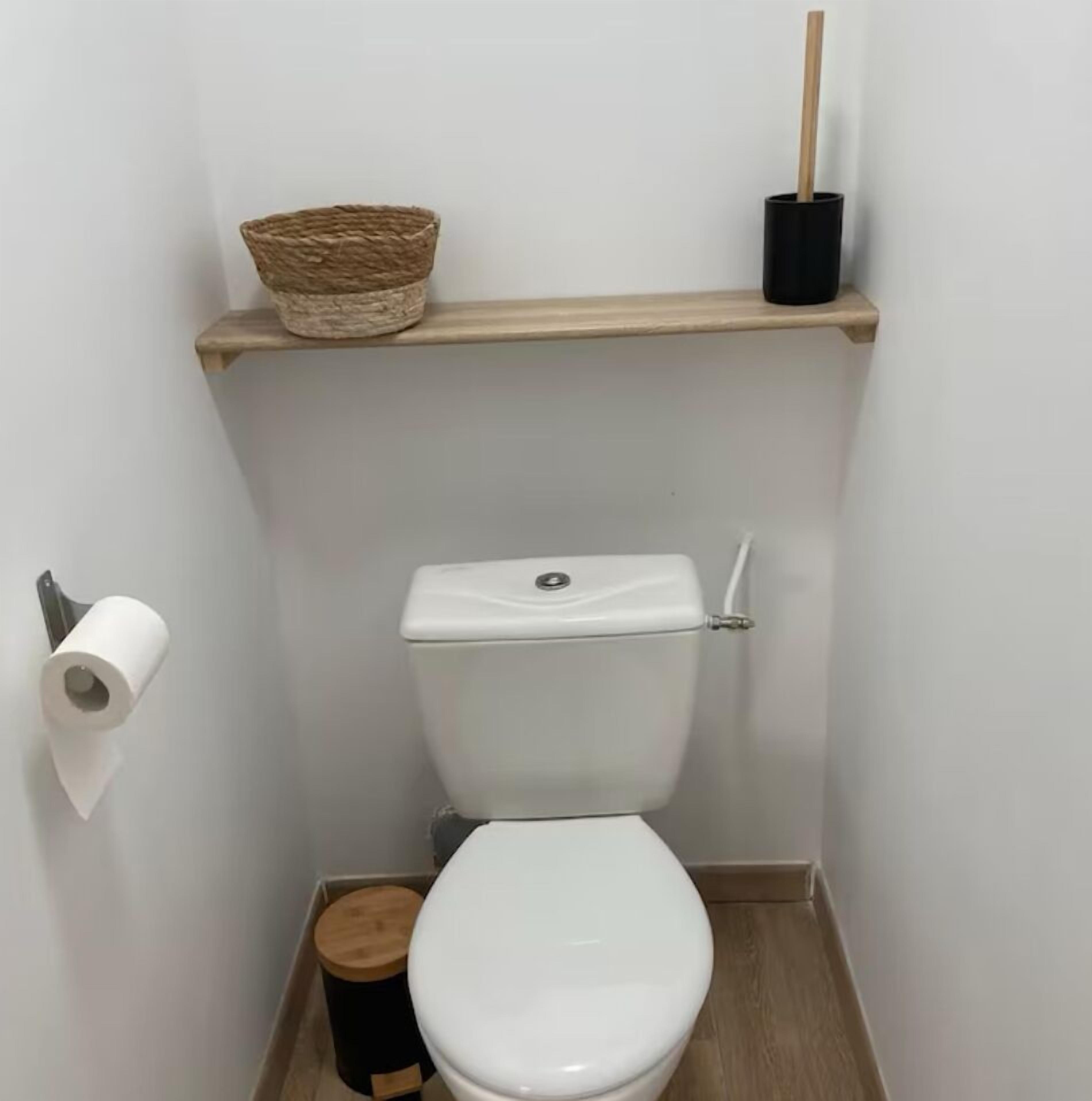 Um banheiro pequeno com vaso sanitário branco, piso de madeira, lixeira preta, rolo de papel higiênico e uma cesta de vime em uma prateleira.