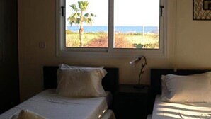 A bedroom with two beds, a window with a view of the beach and palm tree, and a nightstand with a lamp. - Ayia Thekla Villa Poppeli (Ayia Thekla)