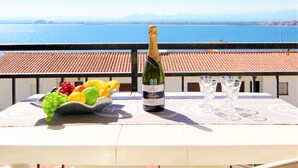 A table with a bottle of wine, glasses, and a bowl of fruit on a balcony overlooking a body of water and mountains. - Apartment in Roses by the Beach & Marina (Roses)