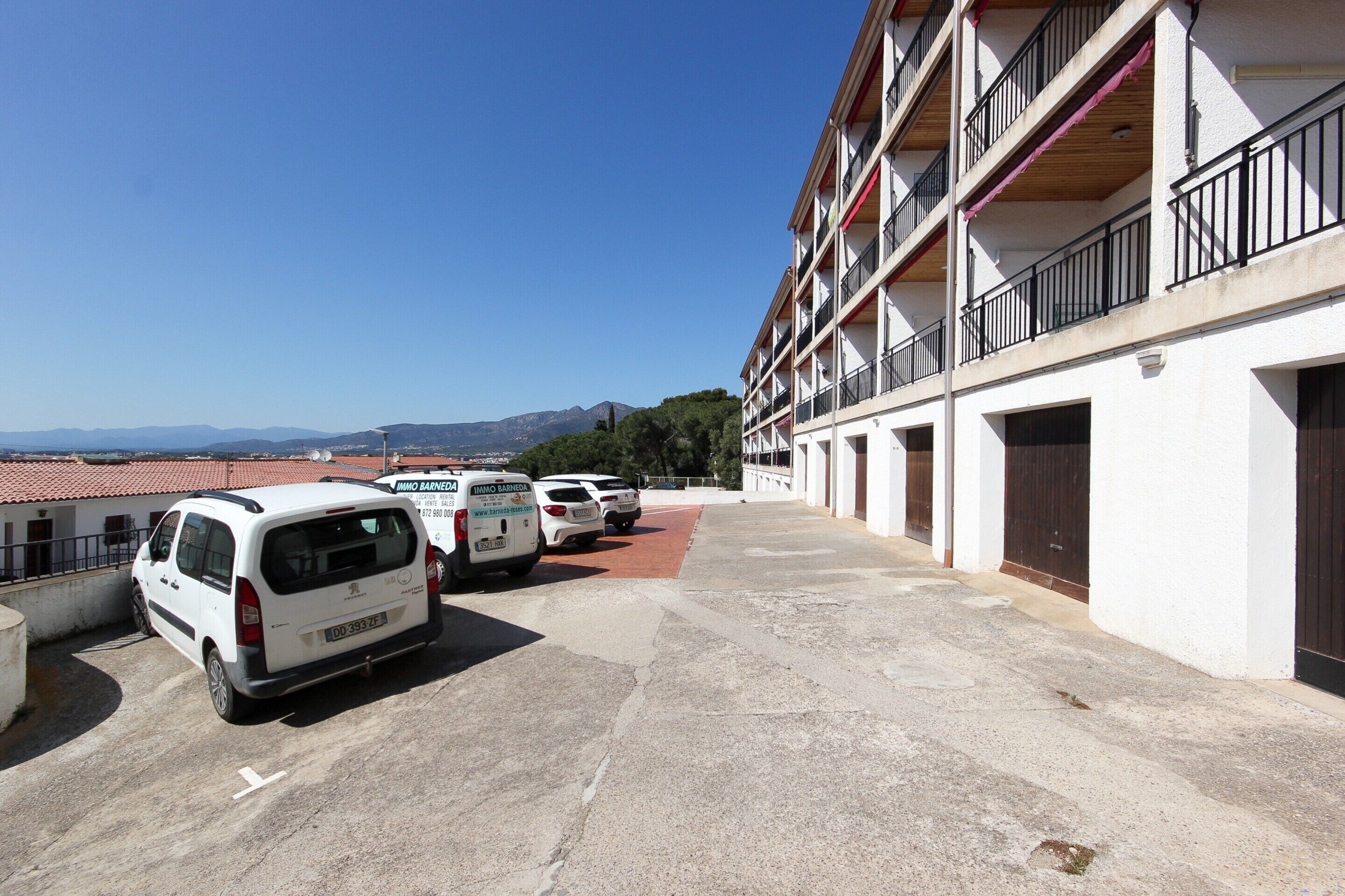 A parking area with several white vans parked in front of a building with multiple balconies and a red-tiled roof.