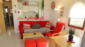A modern living room with a red sofa, matching ottomans, a wooden coffee table, and a dining area with a table and chairs. A kitchen is visible in the background with white cabinets and a clock on the wall. - Apartment by Roses Beach With Pool (Roses)