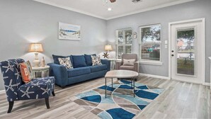 Ein Wohnzimmer mit einem blauen Sofa, einem runden Couchtisch und einem Sessel mit Sternmuster. Es gibt einen Deckenventilator, ein Bild an der Wand und ein Fenster mit Jalousien.
