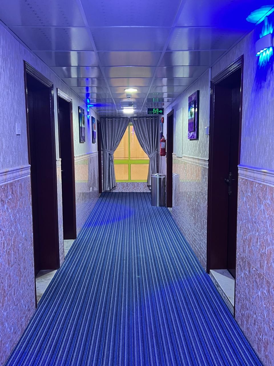 A hotel corridor with a blue-striped carpet, doors on both sides, and a window with curtains at the end.