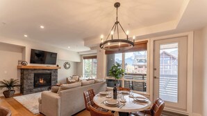 A modern living room with a fireplace, a dining area with a round table and chairs, a large window with a view of buildings, and a cozy sofa. - Top floor 2 level w hot tub - Modern Mountain Oasis (Canmore)