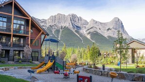 A playground with slides and swings, a building with a balcony, and a mountain backdrop. - Top floor 2 level w hot tub - Modern Mountain Oasis (Canmore)