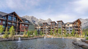 A multi-story hotel with balconies, a central water feature, and mountainous background. - Top floor 2 level w hot tub - Modern Mountain Oasis (Canmore)