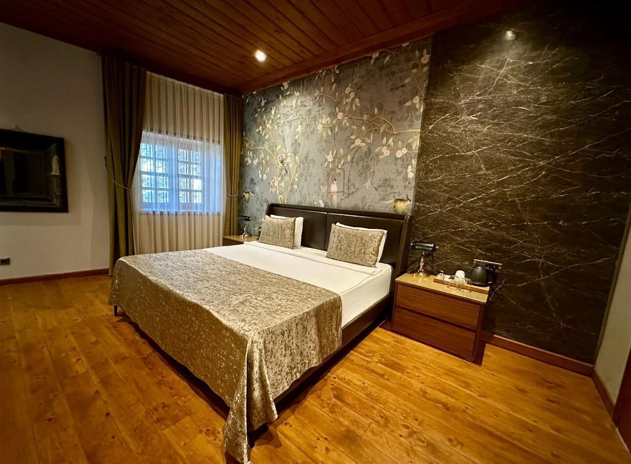 Un dormitorio con cama, suelo de madera, una mesita de noche, pared con estampado floral y ventana con cortinas.