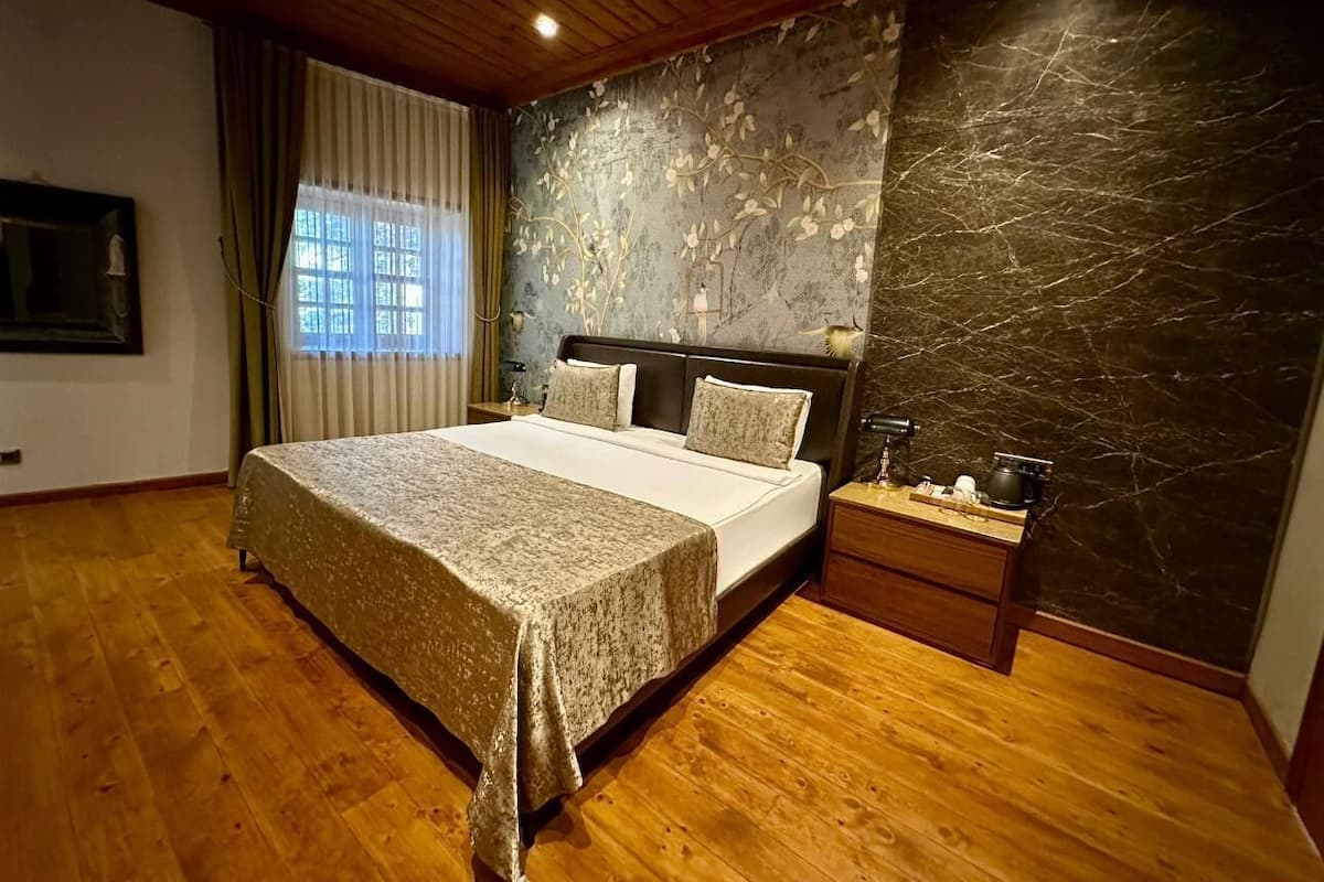 A bedroom with a bed, wooden floor, a nightstand, a wall with a floral pattern, and a window with curtains.