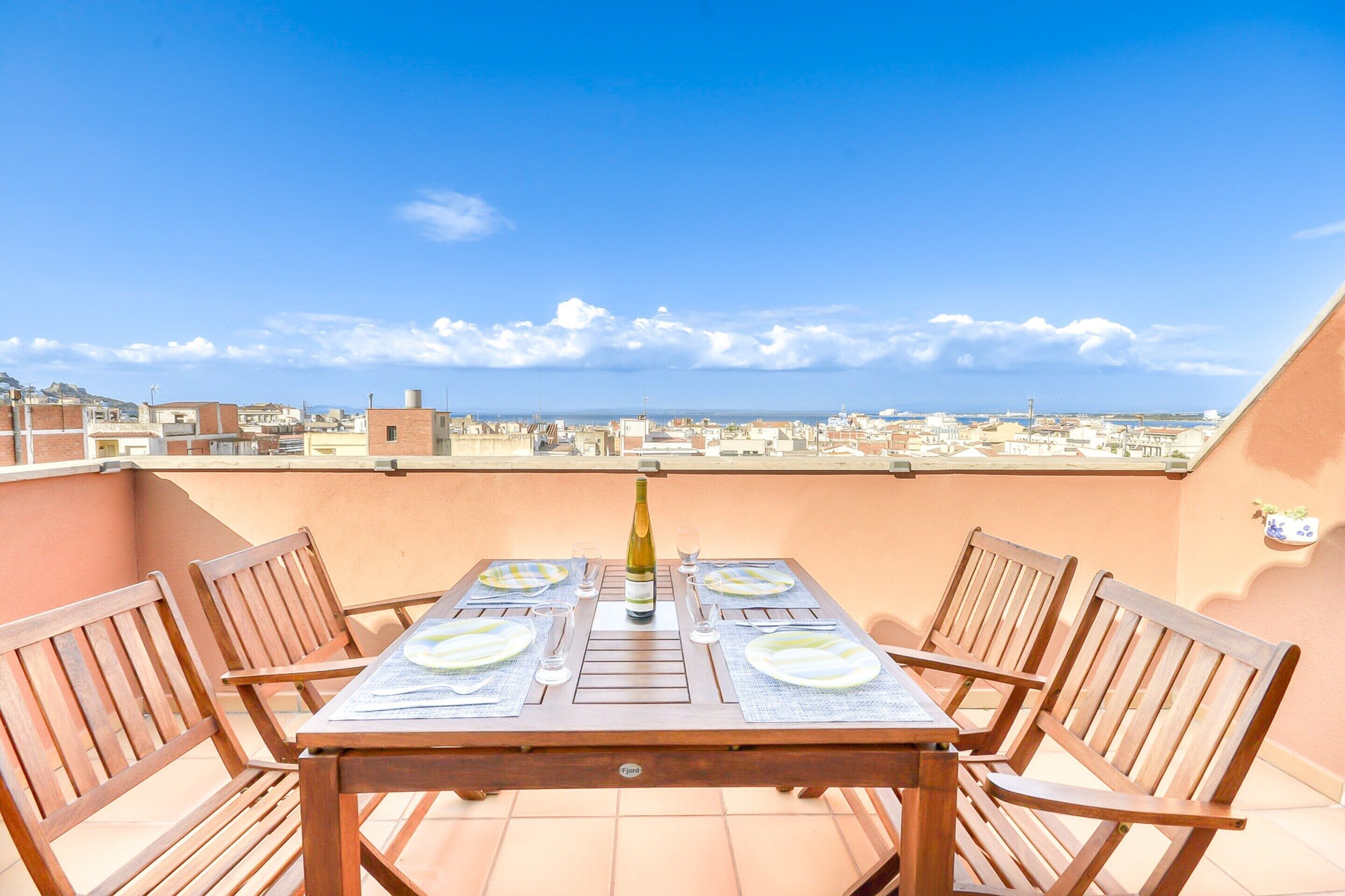 A wooden table set for four with a bottle and glasses, on a balcony with a city view.