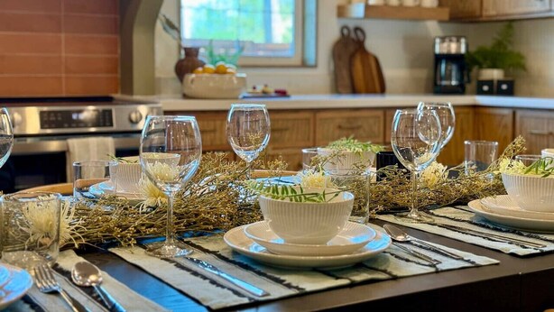 Una mesa de comedor dispuesta con copas de vino, platos y un cuenco de flores, en una cocina moderna con armarios de madera y una ventana.