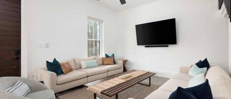 Una sala de estar moderna con ventilador de techo, un televisor de pantalla plana montado en la pared, un sofá color beige con cojines, una mesa de centro y un ventanal con vistas.
