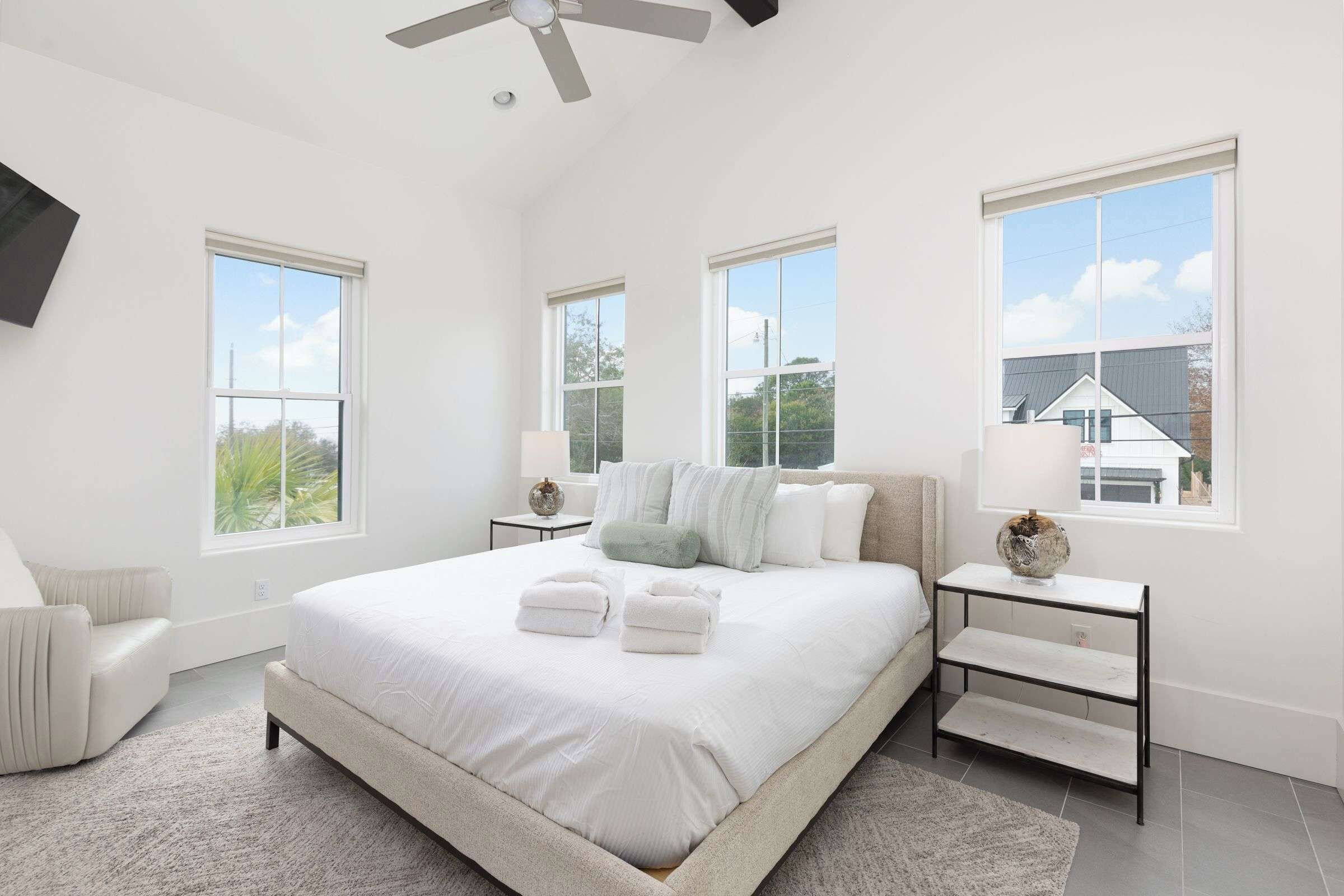A spacious bedroom with a large bed, two bedside tables, a ceiling fan, and multiple windows offering views of the outside.