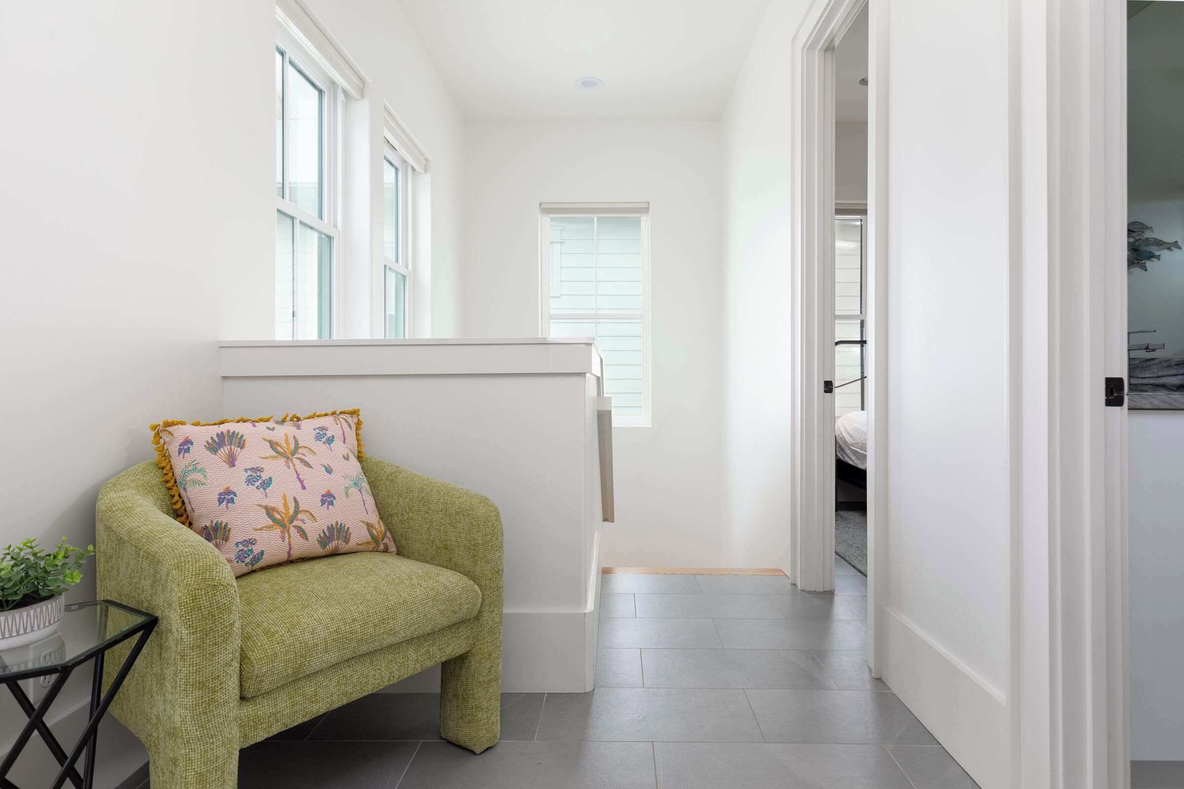 A narrow hallway with a green armchair and a small table with a plant, leading to a bedroom with a bed and a fish wall decoration.