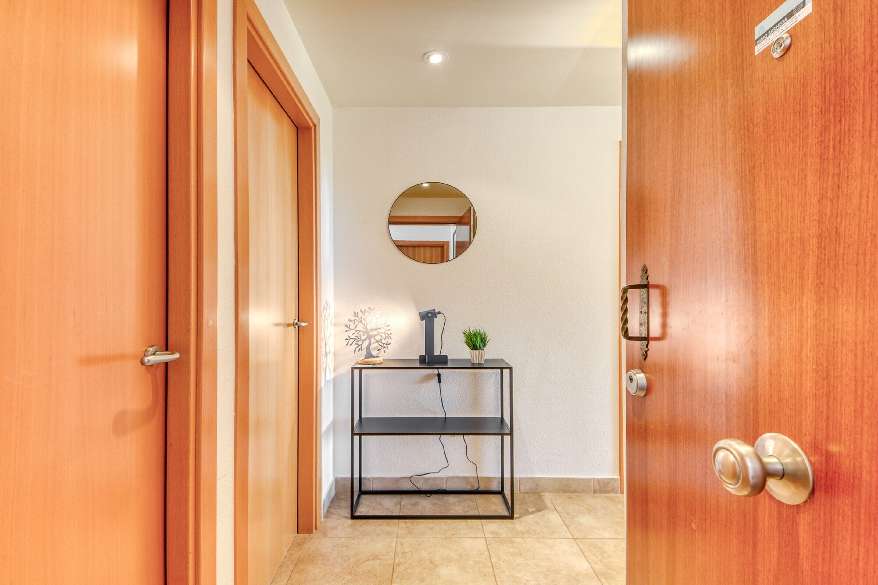Un couloir avec une porte en bois sur la gauche, un miroir rond accroché au mur et une étagère en métal noir sur laquelle sont posés une plante et un petit arbre décoratif.