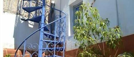 A spiral staircase with ornate metal railings, leading to a balcony with a stone wall backdrop.