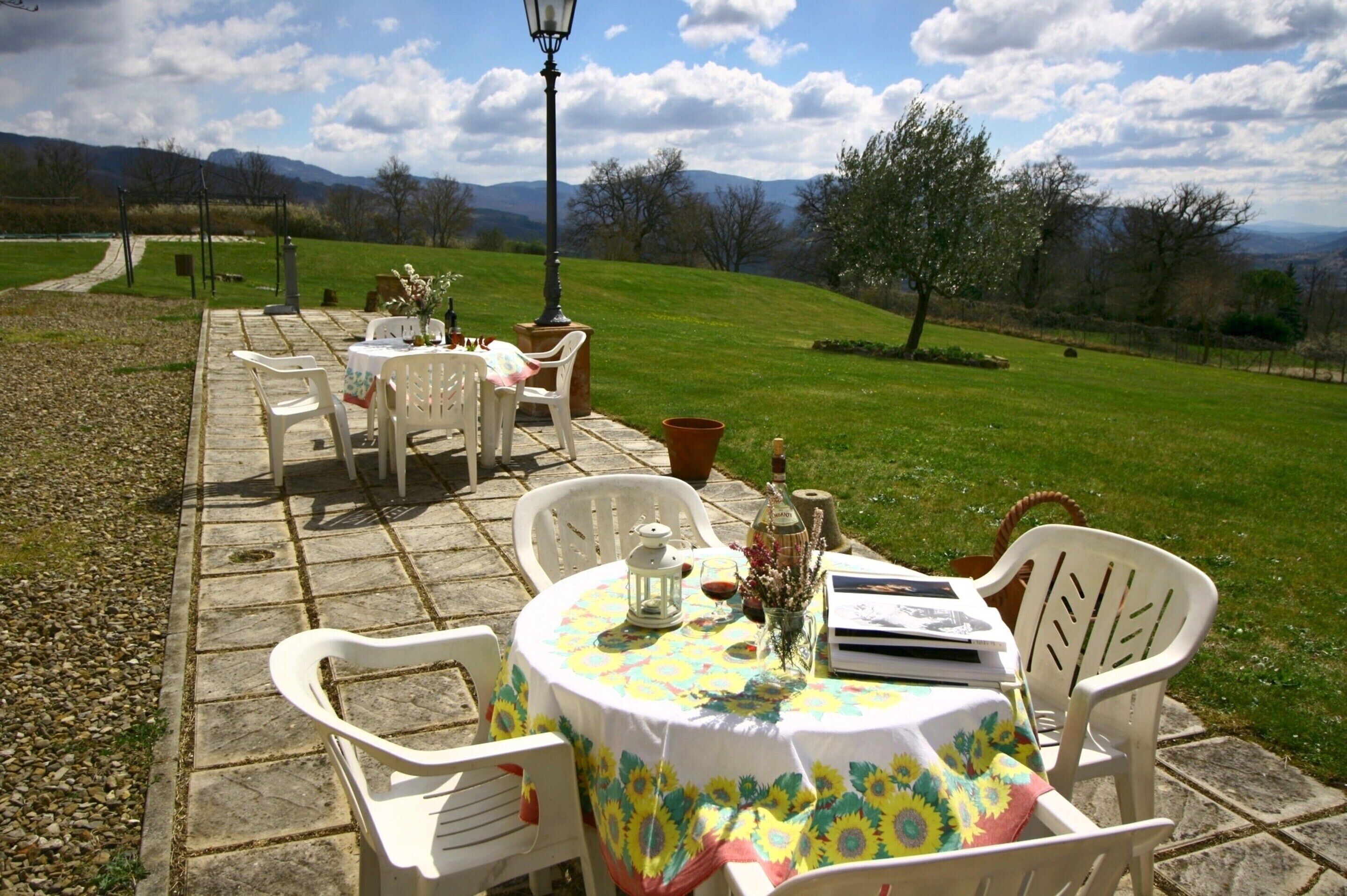 An outdoor patio with stone flooring, white plastic chairs, a round table with a floral tablecloth, and a view of a grassy landscape with trees and mountains in the background.