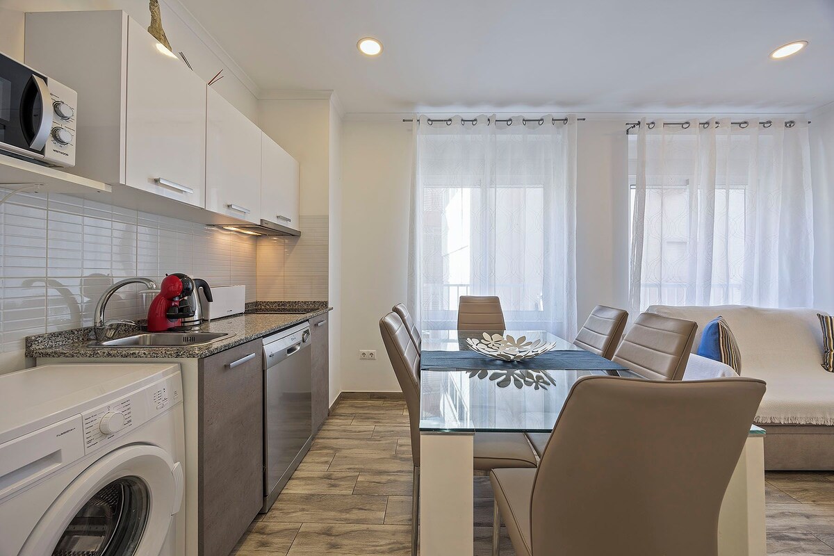A modern kitchen with a built-in washing machine, a coffee maker, and a dining area with a glass table and chairs.