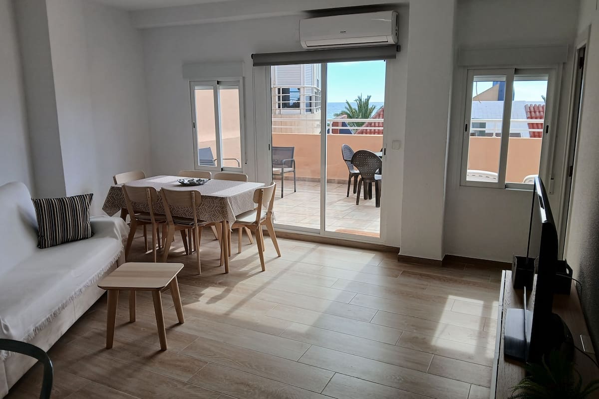 A spacious living area with a dining table, chairs, a sofa, and a sliding glass door leading to a balcony with a view of the sea.